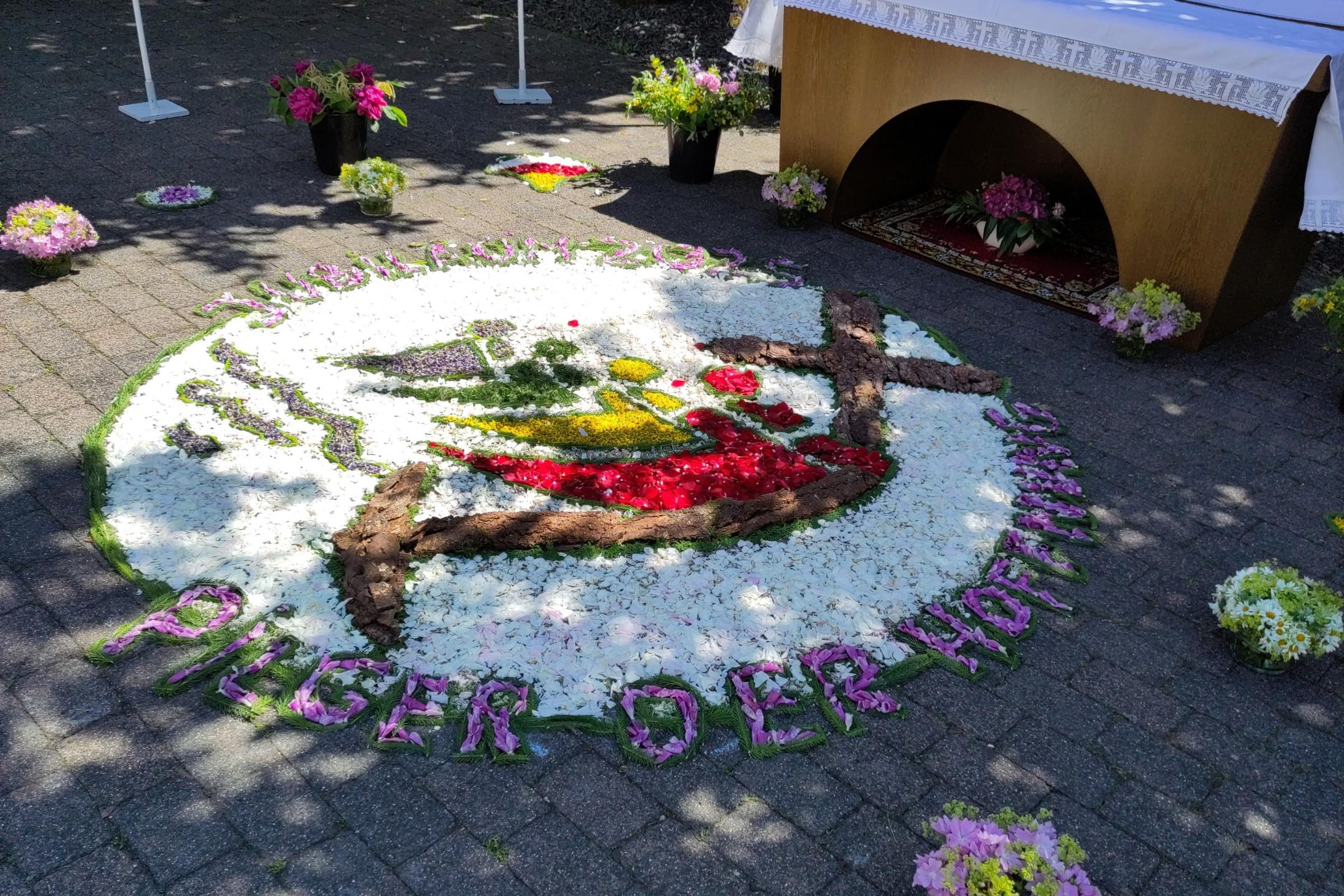 Blumenteppich zur Fronleichnamsprozession auf einem gepflasterten Platz, gestaltet aus bunten Blütenblättern. Im Zentrum des runden Teppichs ist das Logo zum katholischen Jubiläum 2025 mit dem Motto ‚Pilger der Hoffnung‘. Vier stilisierte Personen in Blau, Grün, Gelb und Rot sind nebeneinander dargestellt. Jede Figur besteht aus einem farbigen Kreis als Kopf und einem geschwungenen, gleichfarbigen Segment als Körper. Die Figuren greifen mit ihren Formen ineinander und halten gemeinsam ein großes schwarzes Kreuz, das nach unten in einen Anker übergeht. Unterhalb der Figuren verlaufen stilisierte blaue Wellenlinien. Oben links steht bogenförmig der Text ‚Jubiläum 2025‘, unten rechts der Text ‚Pilger der Hoffnung‘. Umgeben ist das Bild von weißen Blüten mit einem grünen Rand. Am unteren Rand ist aus Blüten der Schriftzug ‚Pilger der Hoffnung‘ gelegt. Im Hintergrund steht ein geschmückter Altar mit weißen Tüchern und Blumenschmuck