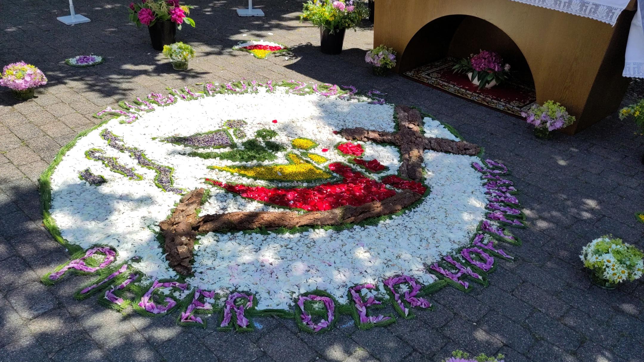 Blumenteppich zur Fronleichnamsprozession auf einem gepflasterten Platz, gestaltet aus bunten Blütenblättern. Im Zentrum des runden Teppichs ist das Logo zum katholischen Jubiläum 2025 mit dem Motto ‚Pilger der Hoffnung‘. Vier stilisierte Personen in Blau, Grün, Gelb und Rot sind nebeneinander dargestellt. Jede Figur besteht aus einem farbigen Kreis als Kopf und einem geschwungenen, gleichfarbigen Segment als Körper. Die Figuren greifen mit ihren Formen ineinander und halten gemeinsam ein großes schwarzes Kreuz, das nach unten in einen Anker übergeht. Unterhalb der Figuren verlaufen stilisierte blaue Wellenlinien. Oben links steht bogenförmig der Text ‚Jubiläum 2025‘, unten rechts der Text ‚Pilger der Hoffnung‘. Umgeben ist das Bild von weißen Blüten mit einem grünen Rand. Am unteren Rand ist aus Blüten der Schriftzug ‚Pilger der Hoffnung‘ gelegt. Im Hintergrund steht ein geschmückter Altar mit weißen Tüchern und Blumenschmuck